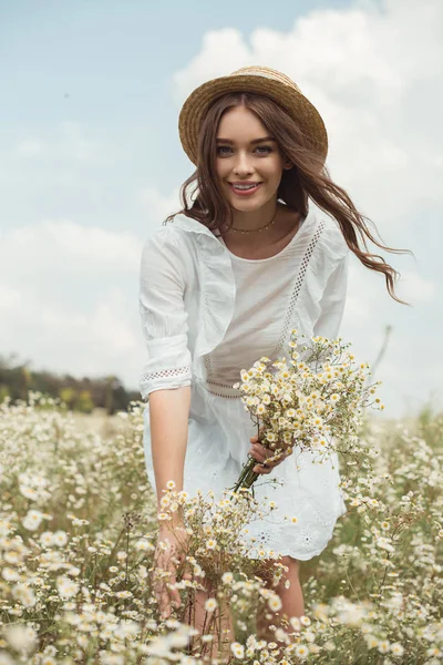 Portrait of pretty woman in white dress with bouquet of wild camomile flowers in field — Stock Photo