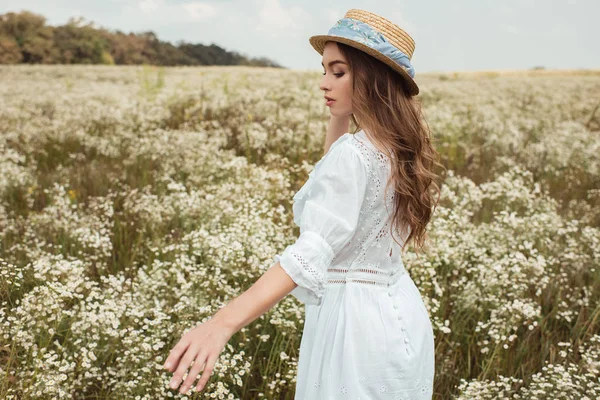 Hübsche nachdenkliche Frau mit Strohhut und weißem Kleid auf einer Wiese mit wilden Blumen — Stockfoto