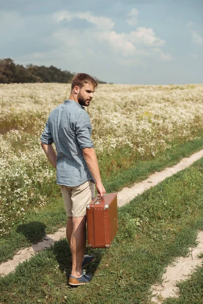 Back view of man with retro suitcase standing in filed with wild flowers on summer day — Stock Photo