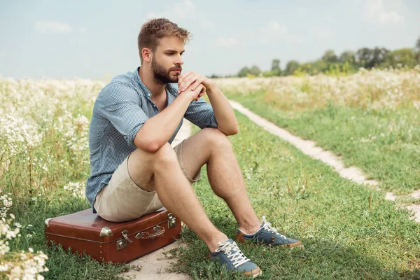 Задумчивый стильный мужчина сидит на ретро чемодан в поле с дикими цветами — стоковое фото