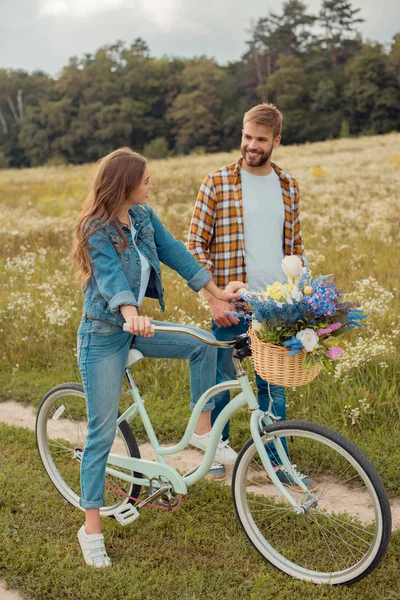 Jeunes amants souriants avec vélo rétro dans un champ avec des fleurs sauvages — Photo de stock