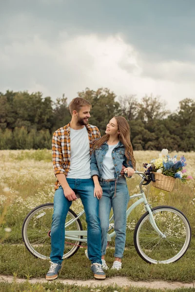 Amantes sonrientes con bicicleta retro mirándose en el campo con flores silvestres - foto de stock