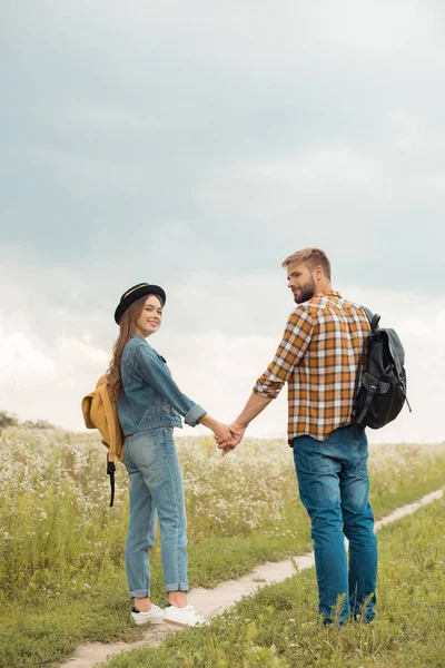 Coppia sorridente con zaini che si tengono per mano nel campo estivo con fiori selvatici — Foto stock