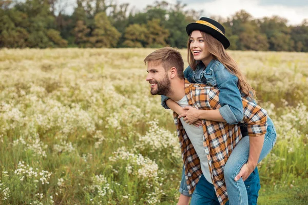 Casal feliz piggybacking juntos no campo de verão com flores selvagens — Fotografia de Stock