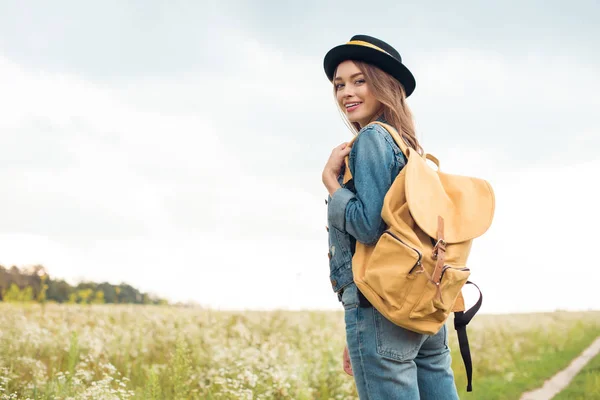 Portrait of young attractive woman in hat with yellow backpack standing in field — Stock Photo