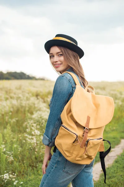Portrait de femme attrayante souriante en chapeau avec sac à dos jaune debout dans le champ — Photo de stock