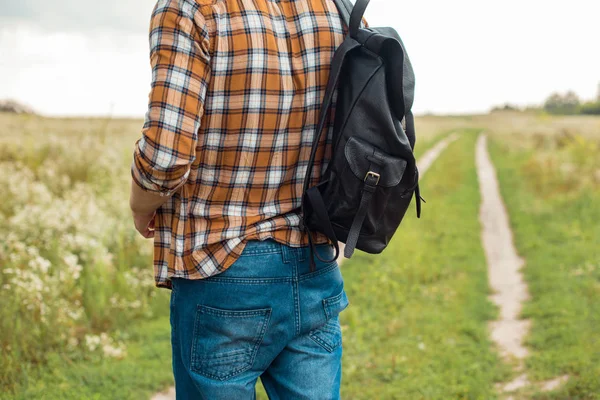 Частичный вид человека в джинсах с черным кожаным рюкзаком, стоящим в поле — стоковое фото