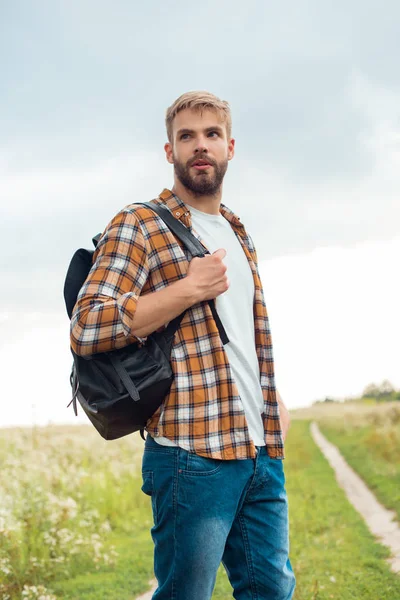 Retrato de hombre pensativo guapo con mochila de cuero negro mirando hacia otro lado en el campo - foto de stock