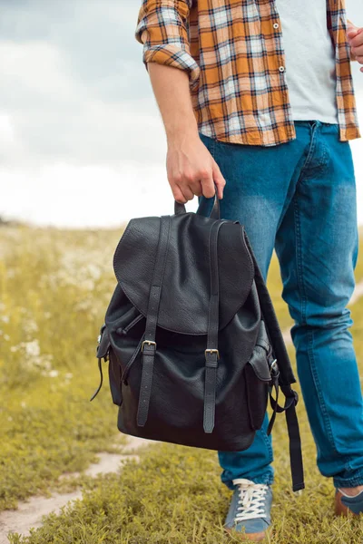 Частичный вид человека в джинсах, держащего черный кожаный рюкзак в поле — стоковое фото