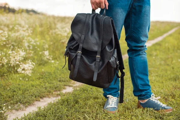 Partial view of man in jeans holding black leather backpack in field — Stock Photo