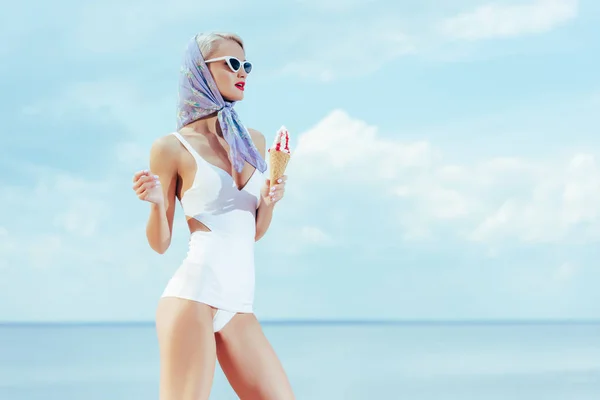 Ragazza alla moda in elegante costume da bagno vintage che tiene il gelato e posa vicino al mare — Foto stock