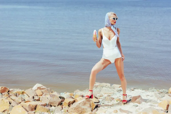 Élégante fille en maillot de bain vintage tenant crème glacée douce et posant sur la plage rocheuse — Photo de stock