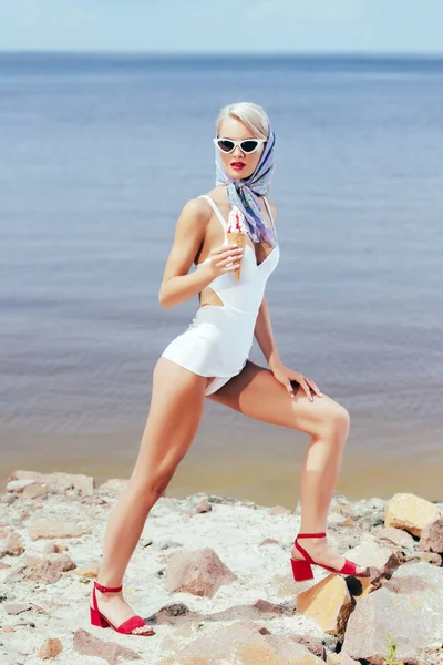 Красива модна дівчина в ретро купальнику тримає морозиво і позує на кам'янистому пляжі — стокове фото