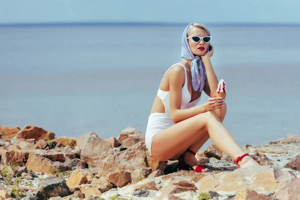 Hermosa mujer en traje de baño vintage celebración de helado y posando en la playa rocosa - foto de stock