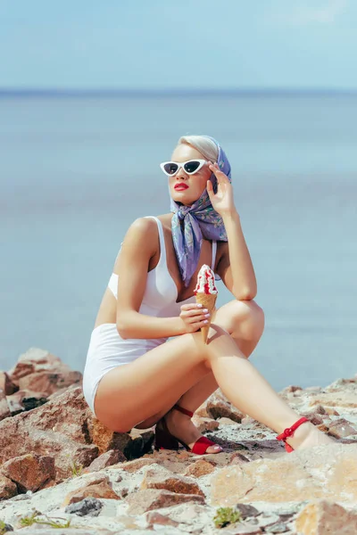 Mujer elegante en traje de baño vintage, bufanda de seda y gafas de sol sosteniendo helado y posando en la playa rocosa - foto de stock