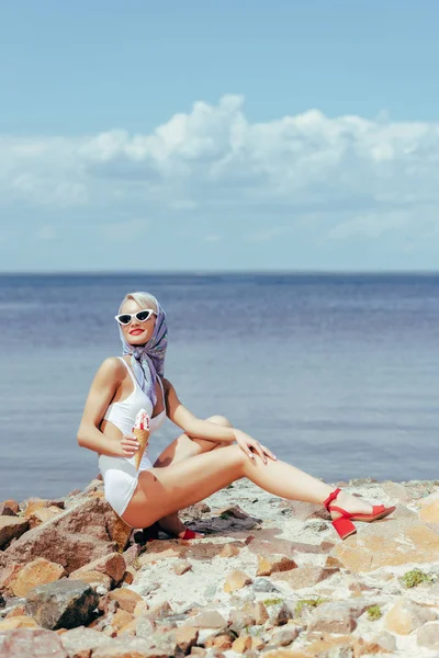 Elegant woman in retro swimsuit holding ice cream and posing on rocky beach at sea — Stock Photo