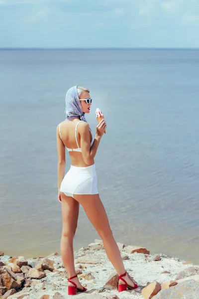 Задний вид элегантной женщины в винтажном купальнике, держащей мороженое и позирующей в море — стоковое фото