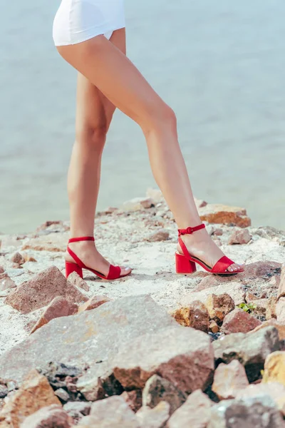Vista ritagliata delle gambe della donna in eleganti sandali con tacco rosso sulla spiaggia rocciosa — Foto stock