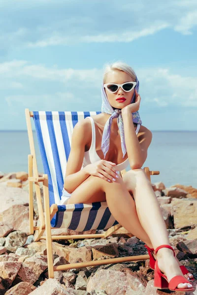 Beautiful girl in stylish sunglasses relaxing in beach chair on rocky shore — Stock Photo