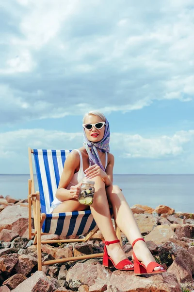 Hermosa chica elegante en gafas de sol sosteniendo tarro con cóctel y relajarse en silla de playa en la orilla rocosa - foto de stock