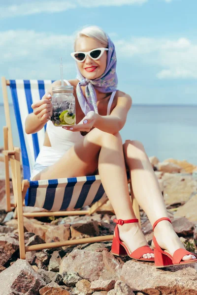 Hermosa chica sonriente sosteniendo limonada en tarro de albañil y descansando en silla de playa - foto de stock