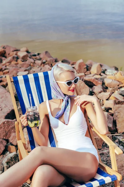 Modische Mädchen halten Glas mit Limonade und entspannen im Strandkorb am felsigen Ufer — Stockfoto