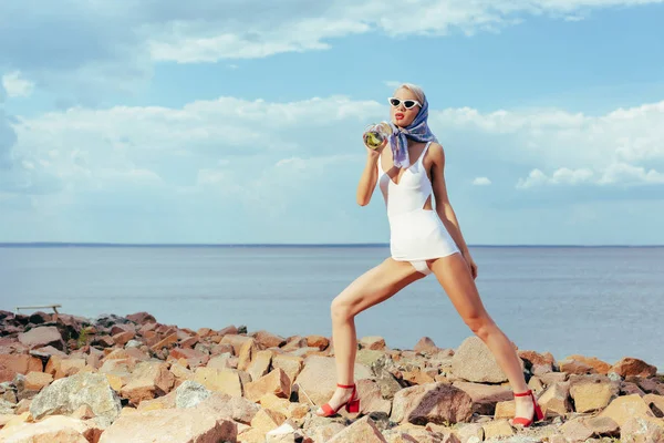 Beautiful girl in white retro swimsuit holding mason jar with fresh lemonade and posing on rocky beach — Stock Photo