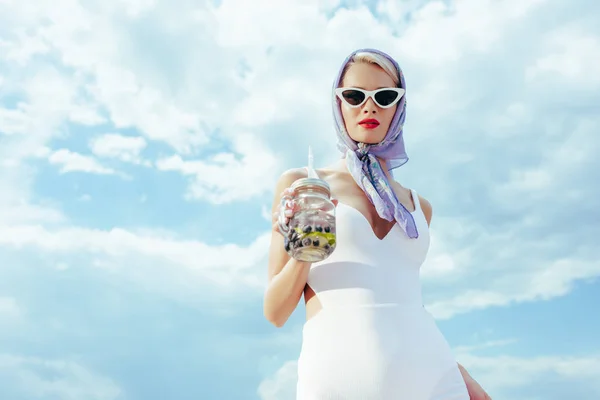 Фешенебельная девушка в солнцезащитных очках и винтажных купальниках держит банку из-под масона со свежим напитком — стоковое фото
