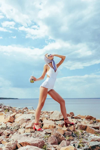Young elegant woman in white vintage swimsuit holding fresh cocktail and posing on rocky beach — Stock Photo