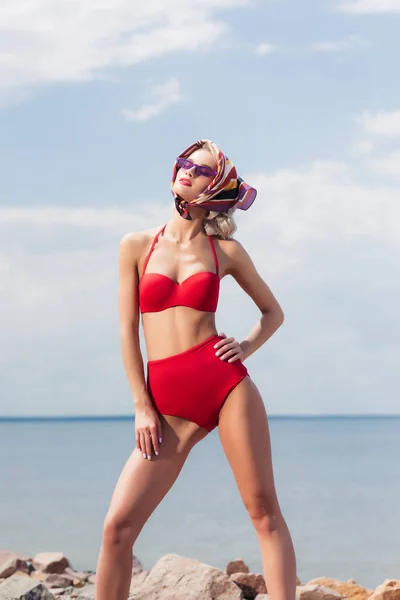 Elegante menina elegante posando em biquíni vermelho, cachecol de seda e óculos de sol na praia rochosa — Fotografia de Stock