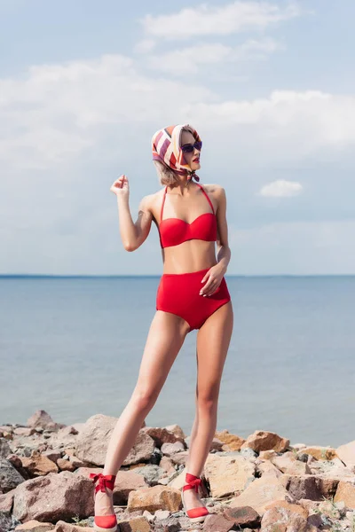 Elegant model posing in red bikini and silk scarf on rocky beach near sea — Stock Photo