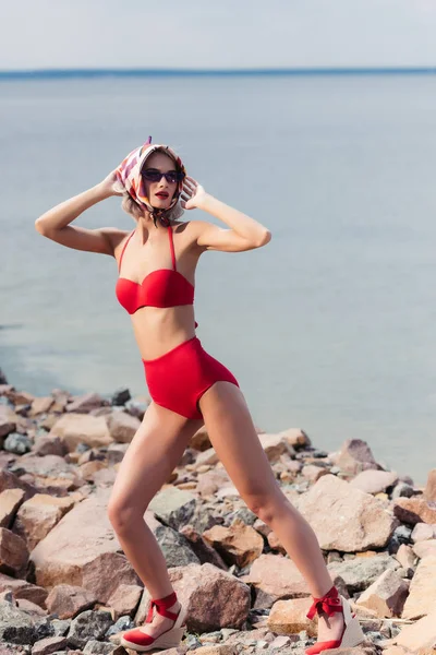 Atractiva chica elegante posando en bufanda de seda y bikini rojo vintage en la playa rocosa - foto de stock