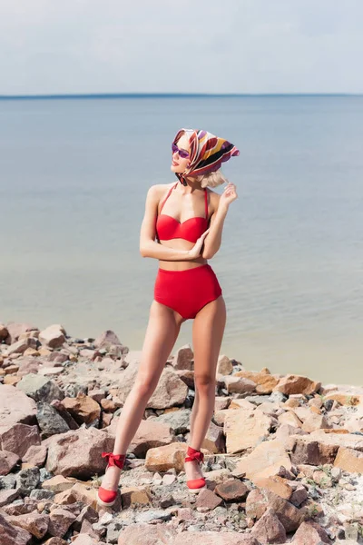 Schönes Mädchen posiert in rotem Bikini und elegantem Schal am felsigen Strand — Stockfoto