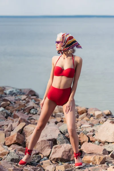 Attractive elegant girl posing in red bikini and silk scarf on rocky beach at sea — Stock Photo