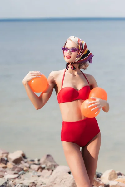 Mulher atraente em biquíni vermelho vintage, óculos de sol e cachecol de seda posando com bolas laranja perto do mar — Fotografia de Stock