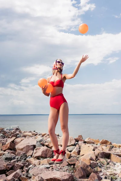 Beautiful stylish woman in red bikini throwing up ball on rocky shore — Stock Photo