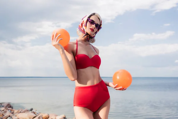 Mujer de moda en bikini rojo, gafas de sol y bufanda de seda posando con bolas en el mar - foto de stock
