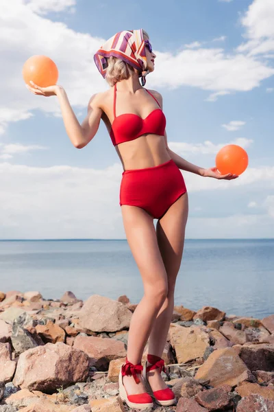 Beautiful elegant girl in red bikini and silk scarf posing with balls on rocky shore — Stock Photo