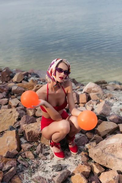 Attractive slim girl in red swimwear, sunglasses and silk scarf posing with balls on rocky beach — Stock Photo