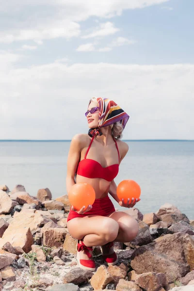 Elegant woman in retro red bikini, sunglasses and silk scarf posing with balls on rocky shore — Stock Photo