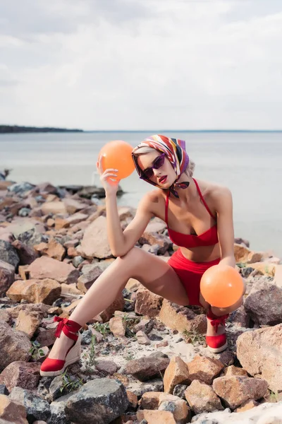 Beautiful stylish woman in red bikini, sunglasses and silk scarf posing with balls on rocky shore — Stock Photo