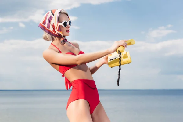 Atractiva mujer en bikini rojo y bufanda de seda posando con teléfono rotatorio amarillo cerca del mar - foto de stock