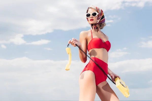 Elegante donna in bikini vintage rosso, occhiali da sole e sciarpa di seta in posa con telefono rotante, cielo nuvoloso sullo sfondo — Foto stock