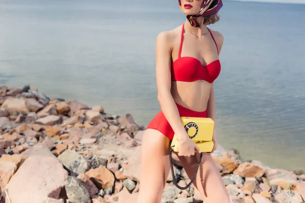 Corte vista do modelo em biquíni vintage vermelho posando com telefone amarelo na praia rochosa — Fotografia de Stock