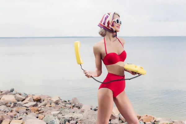 Hermosa mujer joven en bikini rojo y bufanda de seda posando con teléfono rotatorio amarillo cerca del mar - foto de stock