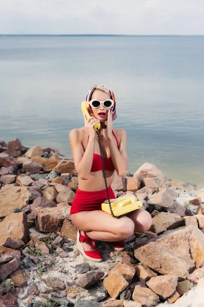 Shocked girl in red bikini and silk scarf talking on vintage rotary telephone — Stock Photo