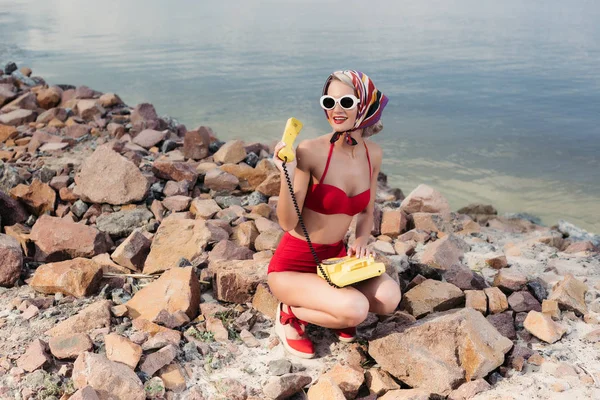 Happy woman in red trendy bikini and silk scarf posing with rotary telephone on rocky beach — Stock Photo