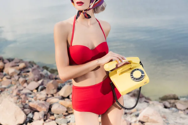 Corte vista do modelo em biquíni retro vermelho posando com telefone rotativo na praia rochosa — Fotografia de Stock