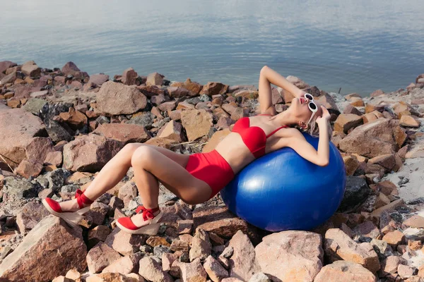Стильна дівчина в старовинному червоному бікіні, розслабляється на синьому м'ячі на кам'янистому пляжі — стокове фото