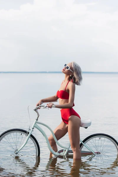 Schöne junge Frau im roten trendigen Bikini sitzt auf dem Fahrrad im Wasser — Stockfoto